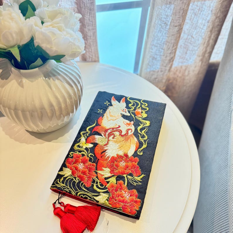 May include: A black embroidered book cover with a fox design in orange and white, set against red flowers and gold detailing. The cover has a red tassel and is on a white table.