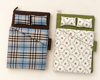 May include: Two miniature beds with fabric bedding. The left bed has a dark brown base, two blue and brown plaid pillows, and a matching plaid blanket. The right bed has an olive green base, two white pillows with small floral prints, and a floral blanket.