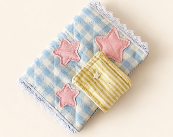 May include: A quilted fabric cover featuring a light blue and white gingham pattern, adorned with three pink fabric stars and a white lace trim. A small yellow and white striped pocket is attached, featuring a tiny white star embellishment.