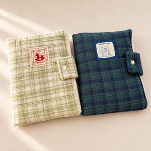 May include: Two rectangular fabric notebook covers with a plaid pattern. The left cover is light green and white with a patch featuring a red rocking horse. The right cover is dark green and navy blue with a patch labeled PRETTY CAT. Both have a star-shaped button closure.