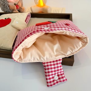 May include: A red and white gingham pouch with a cream-colored interior lining, featuring a red felt carrot appliqu&eacute; on the front flap and a white snap button closure. A second cream-colored quilted pouch with a red apple appliqu&eacute; is visible nearby.