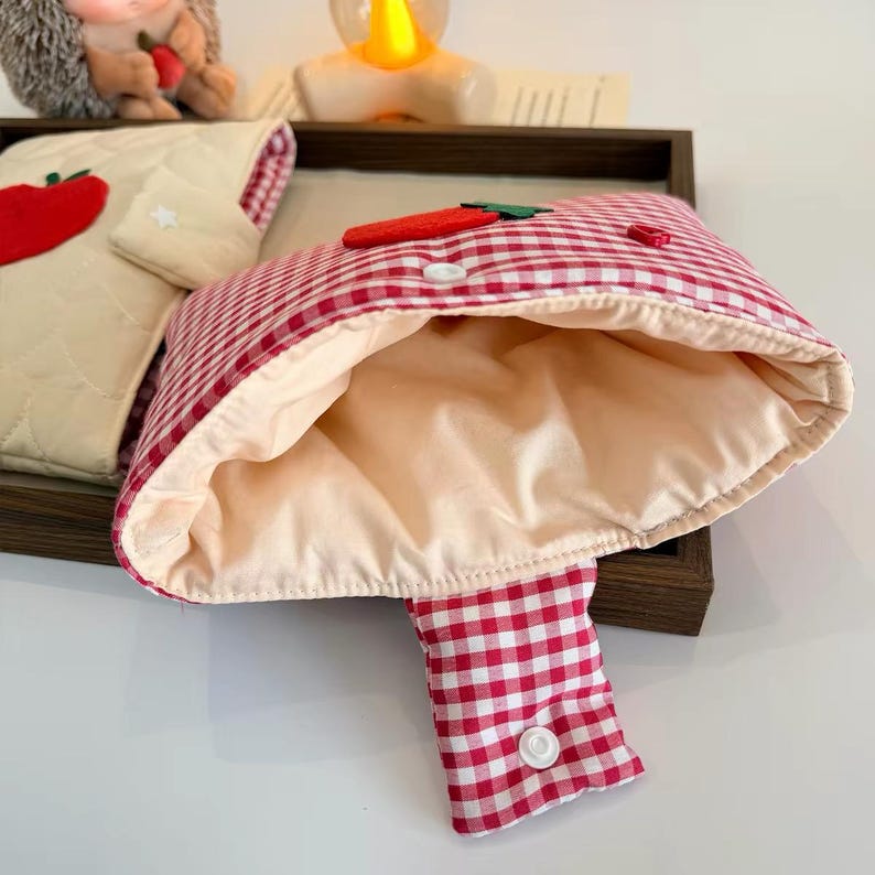 May include: A red and white gingham pouch with a cream-colored interior lining, featuring a red felt carrot appliqu&eacute; on the front flap and a white snap button closure. A second cream-colored quilted pouch with a red apple appliqu&eacute; is visible nearby.