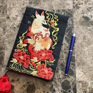 May include: Black embroidered journal cover with a fox design in red and orange, surrounded by red flowers and gold details. A blue pen lies next to the journal. A red tassel hangs from the spine.