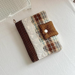 May include: A quilted fabric notebook cover featuring a brown and white polka dot spine, a white lace trim with heart cutouts, and a brown, blue, and beige plaid pattern. A brown polka dot flap with a single white button secures the cover.