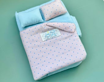 May include: A miniature doll bed set featuring a light blue mattress, a solid light blue pillow, and a light pink pillow with blue polka dots. The matching light pink blanket with blue polka dots has a white label with two blue dolphins.
