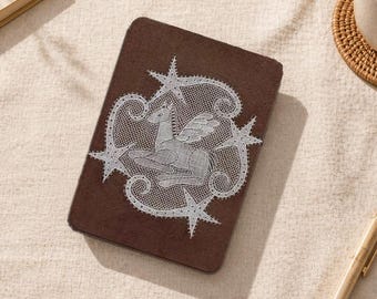 May include: A brown rectangular case with a white lace design of a winged horse and star accents. The case is on a beige fabric, with a pen and a small woven coaster nearby. The case is approximately 15 cm long.