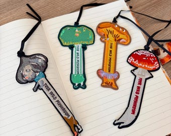 May include: Four embroidered bookmarks with black ribbon ties, each featuring a unique mushroom-inspired character design. Each bookmark includes the text "YOUR WILD MUSHROOM" stitched vertically along the stem, showcasing varied colours like grey, green, orange, and red.