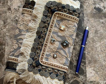 May include: A rectangular clutch bag featuring a beige floral lace overlay on a grey fabric base, adorned with metallic coin-like embellishments, cream-coloured fringe trim, and scattered crystals. A blue ballpoint pen is placed beside the clutch.