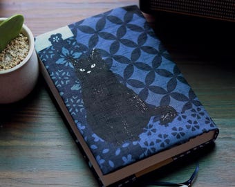 May include: A rectangular notebook with a fabric cover featuring a dark blue and light blue geometric floral pattern. A solid black cat silhouette is printed on the front cover. The notebook includes a thin dark blue ribbon bookmark with a small white flower charm attached to the end.
