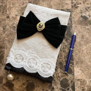 May include: A rectangular white textured notebook featuring a large black velvet bow with a central gold-toned oval brooch depicting a rose. The bottom edge is adorned with white lace trim and a single dangling pearl bead, accompanied by a blue pen.