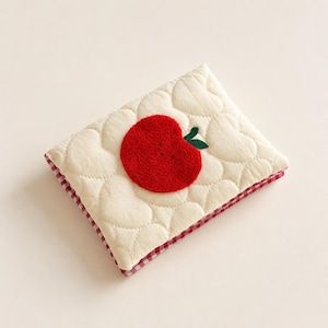 May include: A rectangular quilted pouch featuring an off-white fabric with a heart-shaped stitched pattern. A bright red textured apple appliqu&eacute; with a small green leaf is centered on the front. The edges are trimmed with a red and white gingham fabric.