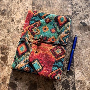 Puede incluir: Un diario rectangular con una cubierta de tela tejida que presenta un patrón geométrico multicolor en tonos rojo, naranja y turquesa. El diario está asegurado con un cordón de cuero marrón y un botón de madera, acompañado de un bolígrafo azul.