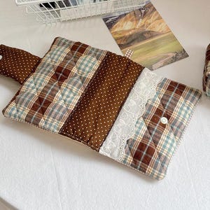 May include: A rectangular quilted fabric pouch featuring a brown and blue plaid pattern, a brown section with white polka dots, and a white lace trim. The pouch includes a flap with a white snap button closure and a matching polka dot tab.