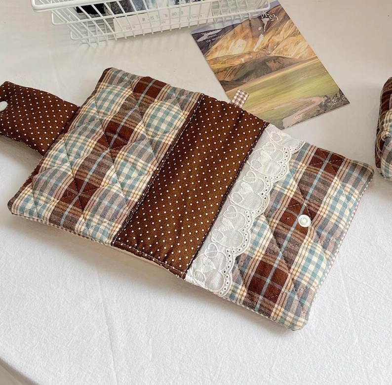 May include: A rectangular quilted fabric pouch featuring a brown and blue plaid pattern, a brown section with white polka dots, and a white lace trim. The pouch includes a flap with a white snap button closure and a matching polka dot tab.