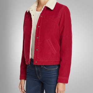 May include: A red corduroy jacket with a cream-colored, fuzzy collar and lining. The jacket has a button-up front, two front pockets, and long sleeves. The model is wearing blue jeans and a white t-shirt underneath.
