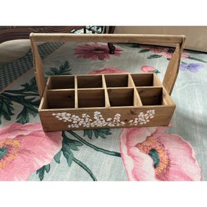 May include: A rectangular wooden caddy featuring eight individual compartments and a tall arched handle. The front panel of the light brown wood is decorated with a white painted floral vine pattern consisting of small leaves and delicate blossoms.