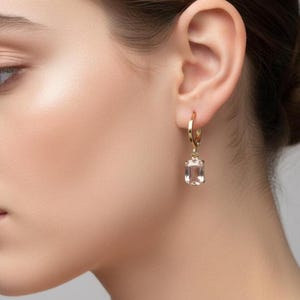 May include: Gold hoop earrings with a rectangular, clear gemstone pendant. The earrings are a simple, elegant design, showcasing the light-coloured gemstone. The earrings are worn on the ear.