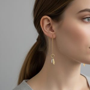 Puede incluir: Pendientes colgantes dorados con un pequeño círculo y dos colgantes de plumas, uno dorado y otro plateado. Los pendientes los lleva una persona con un suéter gris.