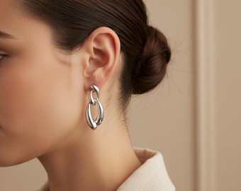 May include: Silver-toned, linked oval earrings. The earrings are a polished, metallic color and have a smooth, rounded shape. The design features two connected oval shapes, creating a modern, minimalist look. These earrings are a jewelry accessory.