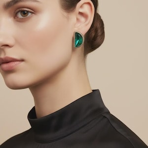 May include: A single earring, half-moon shaped, featuring a large, emerald green gemstone. The earring is set in a gold-coloured metal with a beaded edge. The model wears a black top.