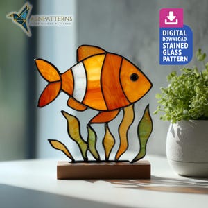 May include: A stained glass fish sculpture with orange, white, and yellow panels, set on a wooden base. The fish has black eyes and is surrounded by green and yellow seaweed. The image includes the text "DIGITAL DOWNLOAD STAINED GLASS PATTERN".