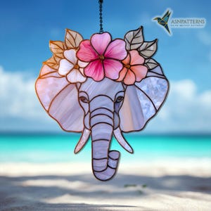 May include: Stained glass elephant with a floral crown, featuring pink and peach flowers, and gray leaves. The elephant is light purple and white, with a chain for hanging. The background shows a beach and blue sky.