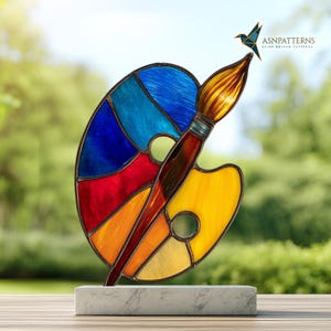 May include: A stained glass art piece featuring a colorful artist's palette and paintbrush. The palette is divided into sections of blue, red, orange, and yellow. The paintbrush has a brown handle and yellow bristles. The artwork is mounted on a white marble base.