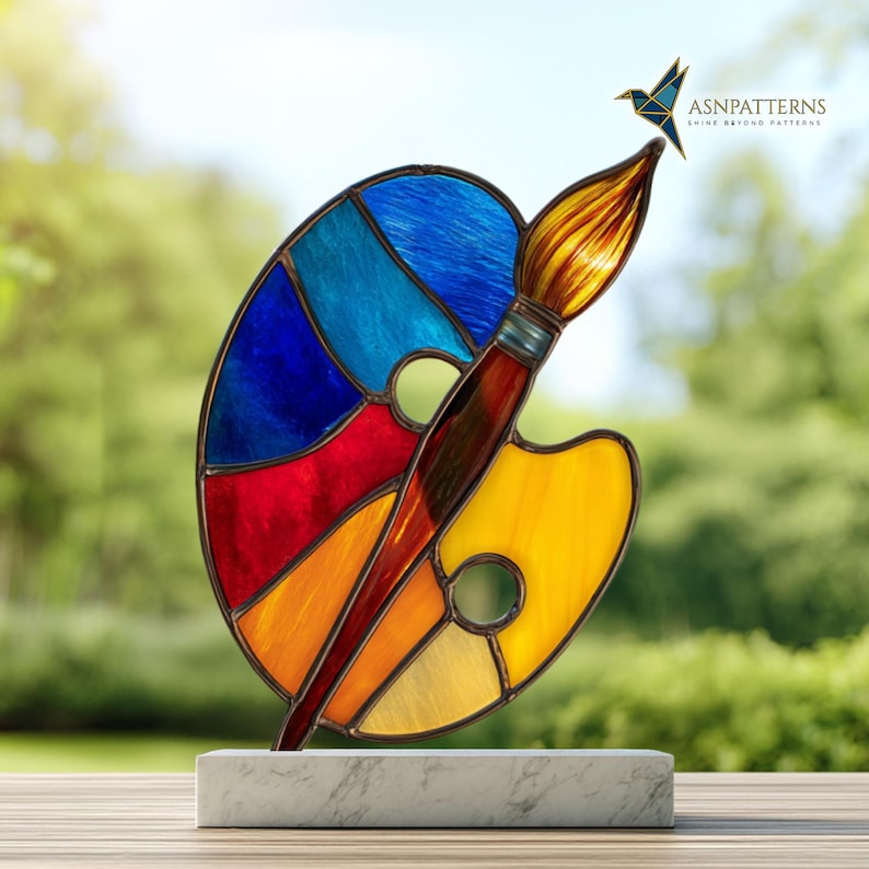 May include: A stained glass art piece featuring a colorful artist's palette and paintbrush. The palette is divided into sections of blue, red, orange, and yellow. The paintbrush has a brown handle and yellow bristles. The artwork is mounted on a white marble base.