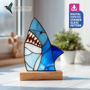 May include: A stained glass shark sculpture in shades of blue, white, and brown, mounted on a wooden base. The shark's mouth is open, revealing teeth. The image includes the text "DIGITAL DOWNLOAD STAINED GLASS PATTERN".