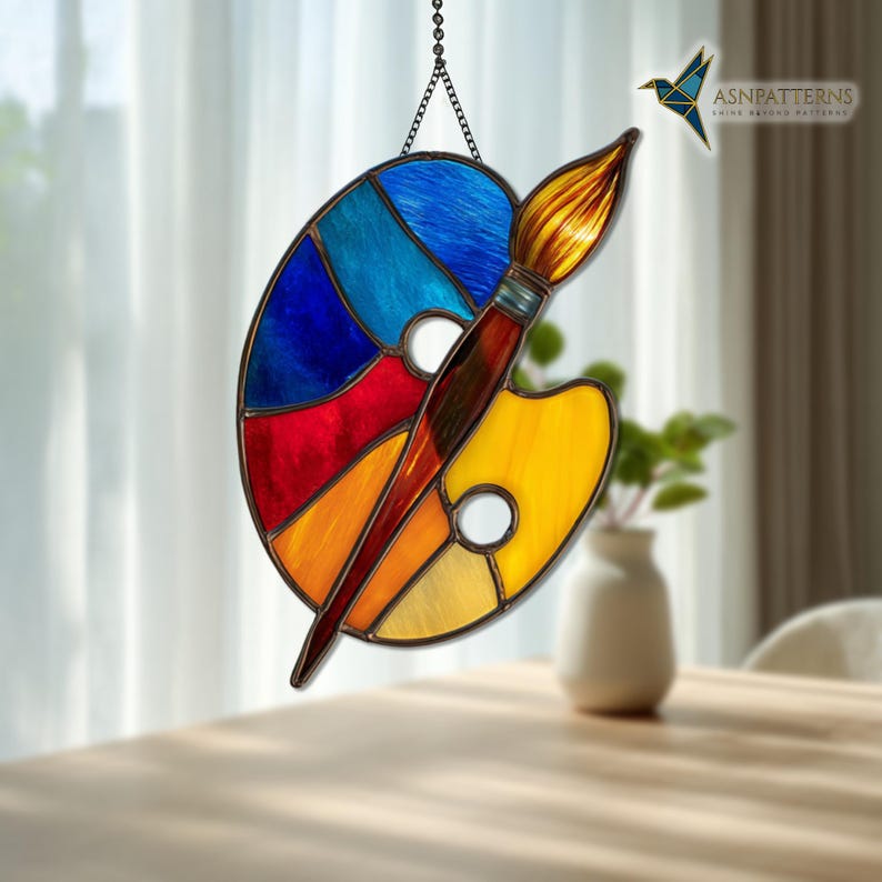 May include: A stained glass art palette with a paintbrush. The palette is divided into sections of blue, red, orange, and yellow. The paintbrush has a brown handle and yellow bristles. The artwork is hanging from a chain.