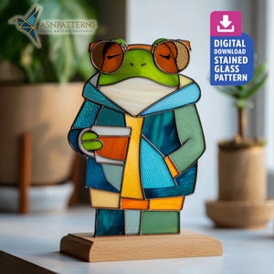 Op de afbeelding: Een gebrandschilderde glazen kikkerfiguur met een zonnebril en een jas, die een koffiekopje vasthoudt. De kikker is groen, blauw en oranje, gemonteerd op een houten voet. De tekst "DIGITAL DOWNLOAD STAINED GLASS PATTERN" is inbegrepen.