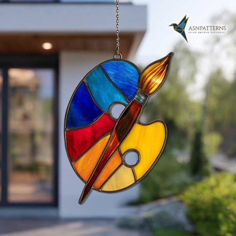 May include: Stained glass art piece depicting an artist's palette and paintbrush. The palette features sections of blue, red, orange, and yellow glass. A brown paintbrush with a golden-yellow bristle is positioned across the palette. The piece is suspended by a chain.