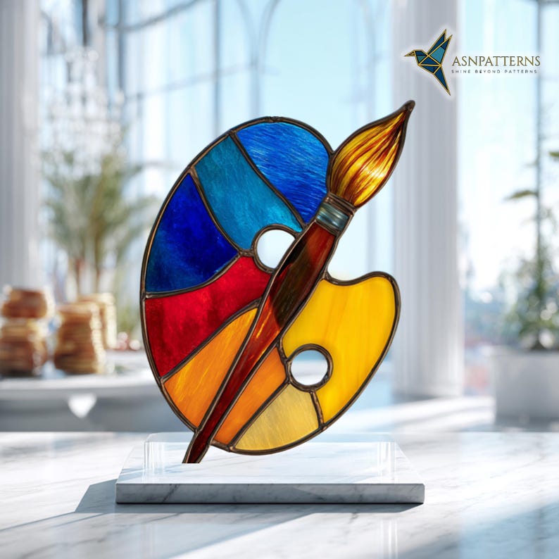 May include: A colorful stained glass art piece depicting an artist's palette and paintbrush. The palette features sections of blue, red, orange, and yellow. The paintbrush is brown with a golden-yellow brush head. The artwork is mounted on a white marble base.