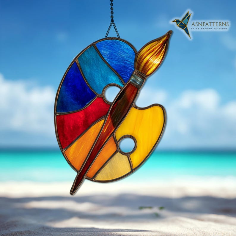 May include: Stained glass art piece featuring a colorful artist's palette with a paintbrush. The palette is divided into sections of blue, red, orange, and yellow. The paintbrush has a brown handle and a golden-yellow brush head. The piece hangs from a chain.