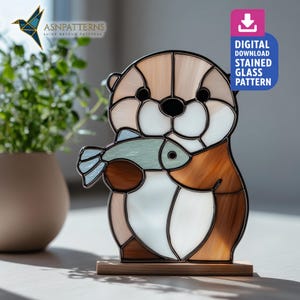 Può includere: Una lontra in vetro colorato che tiene un pesce, con sfumature di marrone, bianco e blu. La lontra è montata su una base di legno. L'immagine include il testo "DIGITAL DOWNLOAD STAINED GLASS PATTERN" e il logo "ASNPatterns".