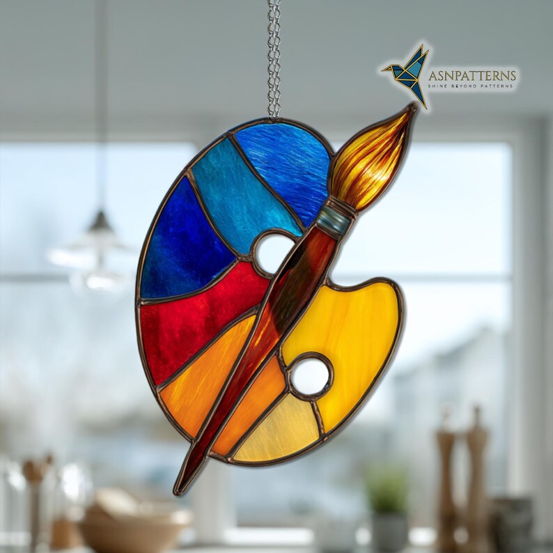 May include: Stained glass art piece featuring a colorful artist's palette and paintbrush. The palette is divided into sections of blue, red, orange, and yellow. A brown paintbrush is positioned across the palette. A small, blue origami hummingbird is above the brush.