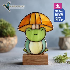 May include: A stained glass frog figurine with a yellow and orange umbrella-like top. The frog is green and blue, mounted on a wooden base. The image includes the text "DIGITAL DOWNLOAD STAINED GLASS PATTERN".