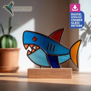 May include: A stained glass shark sculpture in shades of blue, red, and orange, mounted on a wooden base. The shark features sharp teeth and a detailed fin design. The image also includes text that reads "DIGITAL DOWNLOAD STAINED GLASS PATTERN".