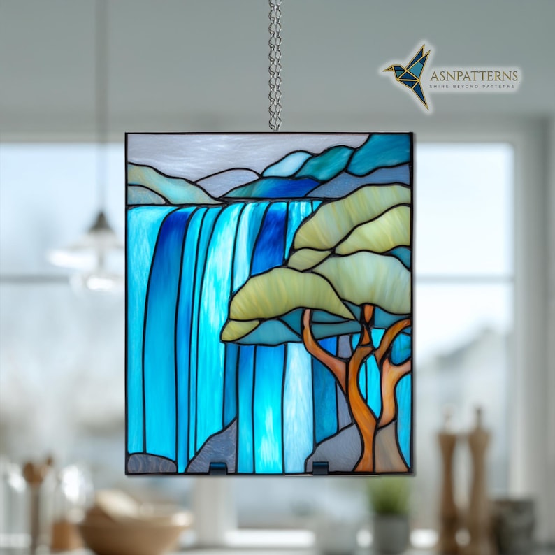 May include: Stained glass panel showing a waterfall scene with blue and turquoise water. A tree with yellow-green leaves and an orange trunk is on the right. The panel is framed in black and hangs from a silver chain.