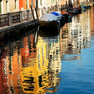 Peut inclure: Un canal à Venise avec plusieurs bateaux amarrés munis de bâches blanches et bleues. L'eau reflète les façades jaune et orange vives des bâtiments adjacents, créant une scène colorée avec une rambarde métallique le long du quai en pierre.