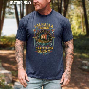 May include: Heather navy t-shirt with a Viking ship graphic. The design includes the text "VALHALLA FORGE CRAFTED FOR GLORY" in a circular emblem with axes and swords. The shirt is a crew neck and has short sleeves.