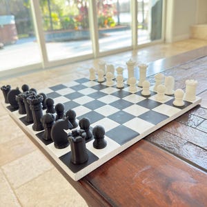 Puede incluir: Un tablero de ajedrez cuadrado con una cuadrícula de casillas alternas blancas y negras. El tablero está dispuesto con un juego completo de piezas de ajedrez blancas y negras, incluyendo reyes, reinas, torres, alfiles, caballos y peones.