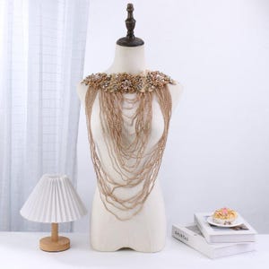 May include: A gold-toned beaded shoulder piece with floral embellishments and cascading chains. The piece is displayed on a mannequin, showcasing its intricate design and draping effect. The shoulder piece is a statement accessory.