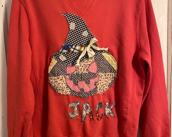 Op de afbeelding: Koraalrood sweatshirt met een patchwork pompoenontwerp. De pompoen heeft een zwart-wit geruite hoed, bloemen en roze accenten en de naam "JACK" in kleurrijke letters. De sweatshirt heeft een ronde hals en lange mouwen.