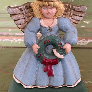 Op de afbeelding: Een keramieken engelenbeeldje met een lichtblauwe jurk en blond haar. De engel houdt een groene krans met rode bessen en een rode strik vast. De vleugels zijn getextureerd bruin van kleur en de basis is groen.