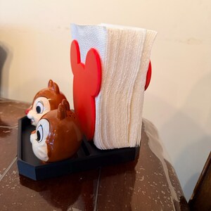May include: A black tray holds a white napkin holder with red Mickey Mouse ears and two Chip and Dale salt and pepper shakers. The shakers are brown with white and black details.