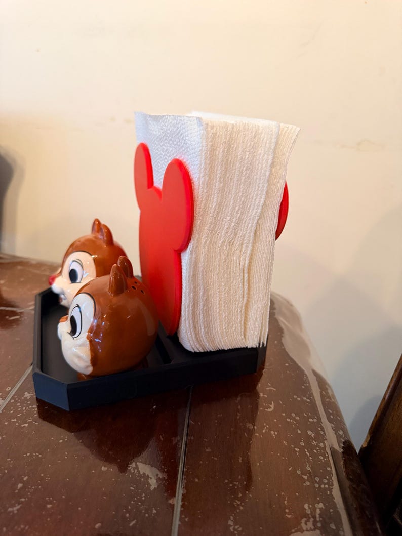 May include: A black tray holds a white napkin holder with red Mickey Mouse ears and two Chip and Dale salt and pepper shakers. The shakers are brown with white and black details.