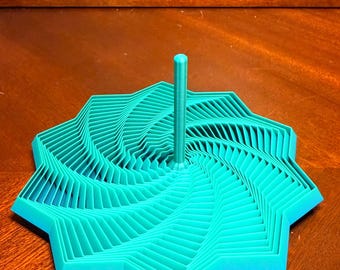 May include: A teal-colored, geometric, 3D-printed object with a central vertical post. The design features a layered, star-like shape with a spiral pattern. The object is made of plastic and sits on a wooden surface.