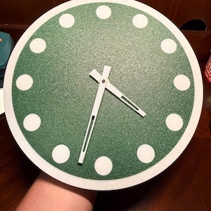 May include: A round wall clock with a green face and white hour markers. The clock hands are white with black accents. The clock's face is surrounded by a white border. The clock is a decorative item.