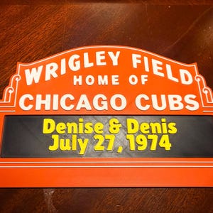 Op de afbeelding: Een rechthoekig bord in oranje, zwart en wit, met bovenaan de tekst "WRIGLEY FIELD HOME OF CHICAGO CUBS". Daaronder staat in een zwarte rechthoek "Denise & Denis July 27, 1974" in geel. Het bord heeft een decoratieve rand.